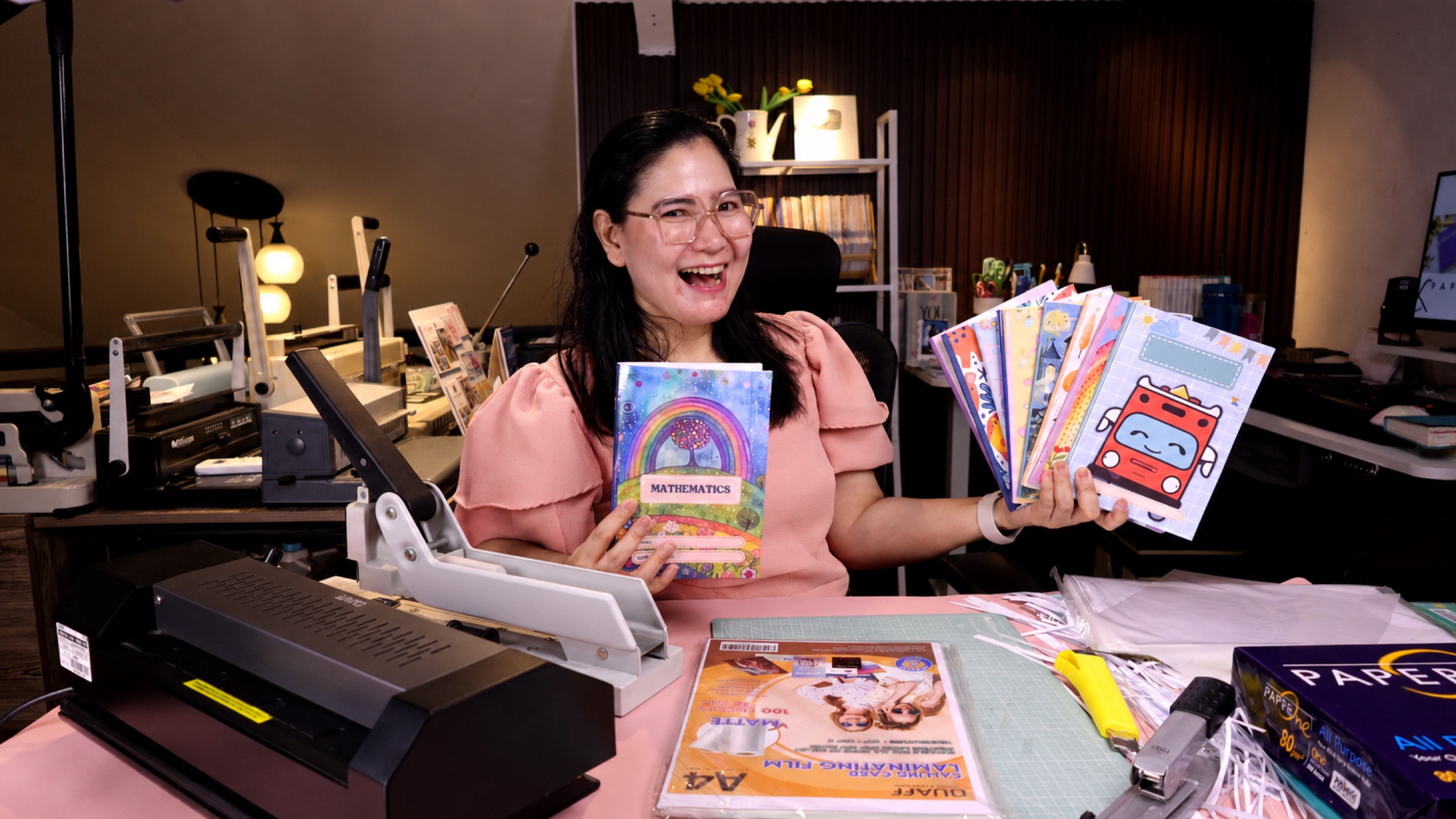 Grace, founder of Print Gracefully, in her home workshop holding handmade notebooks beside her binding equipment